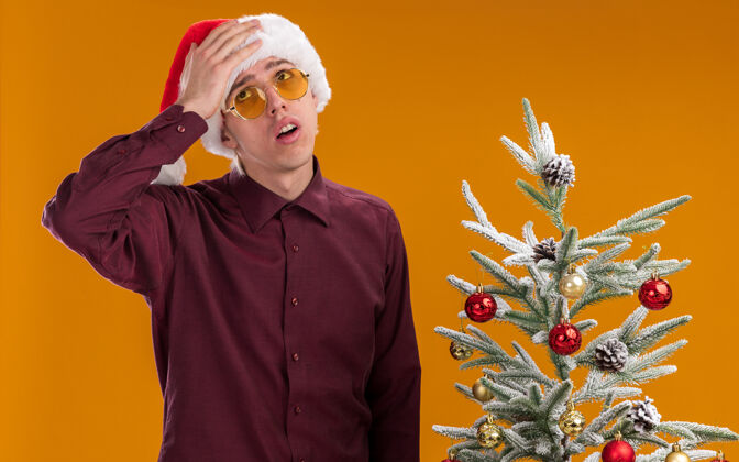 看遗憾的是 年轻的金发男子戴着圣诞帽 戴着眼镜 站在装饰好的圣诞树旁 抬起头来 手放在头上 在橙色的背景下与世隔绝眼镜金发年轻