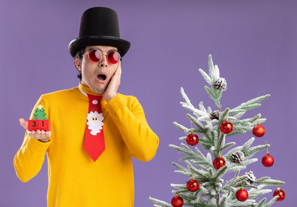 男人一个穿着黄色高领毛衣 戴着黑帽子和滑稽领带的年轻人拿着新年约会的方块 惊讶地站在紫色背景下的圣诞树旁看着相机相机立方体圣诞节