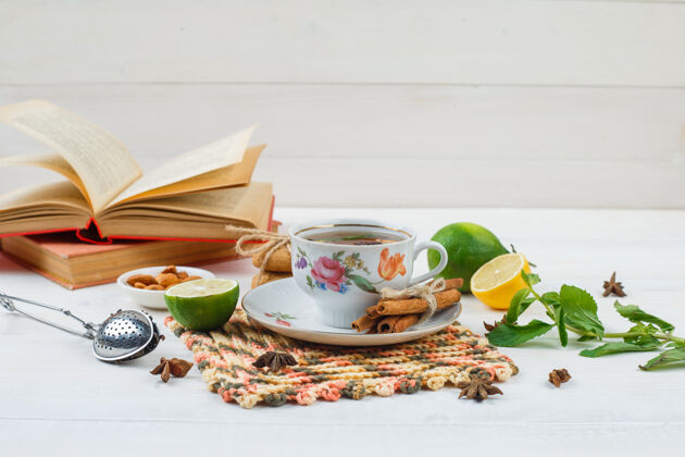碗一杯肉桂柠檬茶放在方形的柠檬垫上 一碗杏仁 滤茶器和书放在白色的表面上薄荷角草药