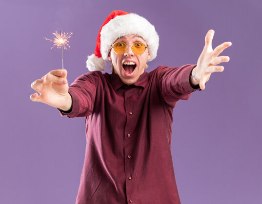 帽子令人印象深刻的年轻金发男子戴圣诞帽和眼镜伸出节日火花和手做欢迎手势孤立在紫色的墙壁上欢迎年轻印象深刻