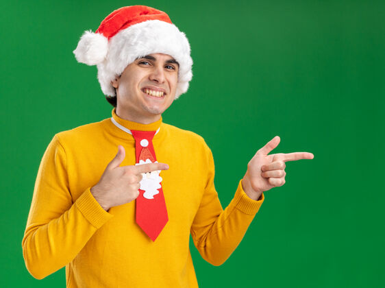 领带一个穿着黄色高领毛衣 戴着圣诞帽 打着滑稽领带的年轻人看着摄像机 兴高采烈地笑着 用食指指着站在绿色背景上的一边男人搞笑指指点点