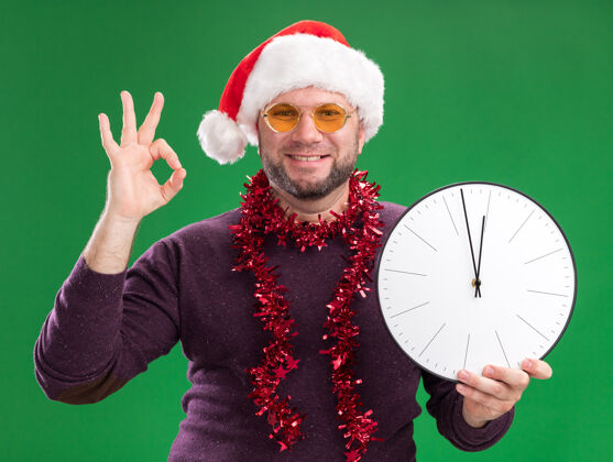 脖子微笑着的中年男子戴着圣诞帽 脖子上戴着金箔花环 戴着眼镜 手里拿着时钟 绿色墙上孤立着一个“一切正常”的牌子时钟帽子戴着