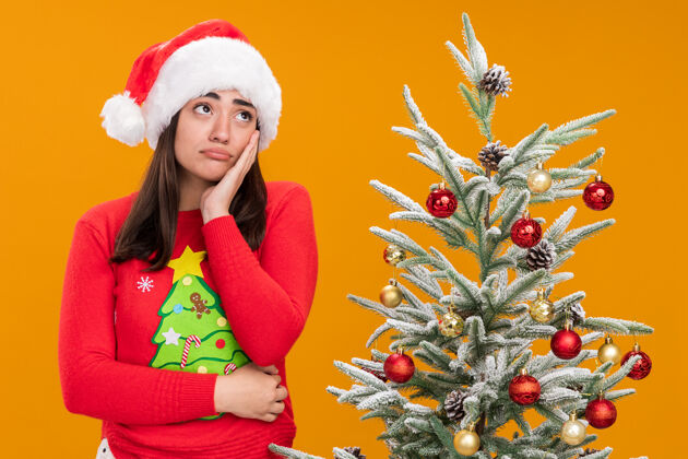 悲伤戴着圣诞帽的悲伤的白人女孩把手放在脸上 看着侧面站立看树
