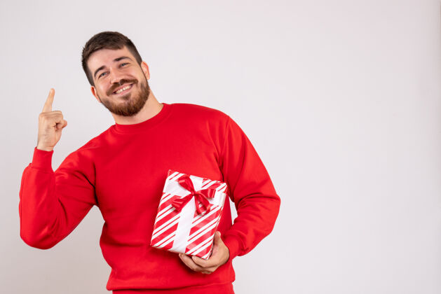 英俊身穿红衫的年轻人手持圣诞礼物 微笑着站在白墙上男人男人成人