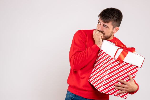 英俊身穿红衫的年轻人在白色墙上的盒子里拿着圣诞礼物持有男性年轻