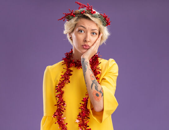 女人焦急的金发女郎头戴圣诞花环 脖子上戴着金箔花环 手放在脸上 在紫色的墙壁上看起来很孤立 有复制空间保管花环圣诞节