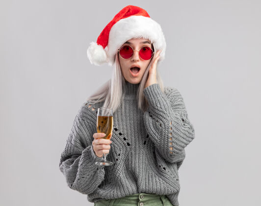 年轻的年轻的金发女郎穿着冬天的毛衣 戴着圣诞帽 手里拿着一杯香槟 看上去很惊讶毛衣圣诞老人冬天