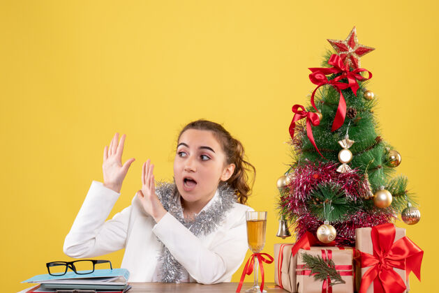 坐前视图：女医生坐在桌子后面 拿着圣诞礼物和黄色背景上的树礼物前面护士