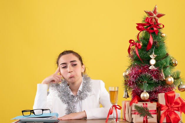 插花正面图：坐在桌子后面的女医生 带着圣诞树和礼盒 在黄色的背景下 她感到悲伤前面女性女医生