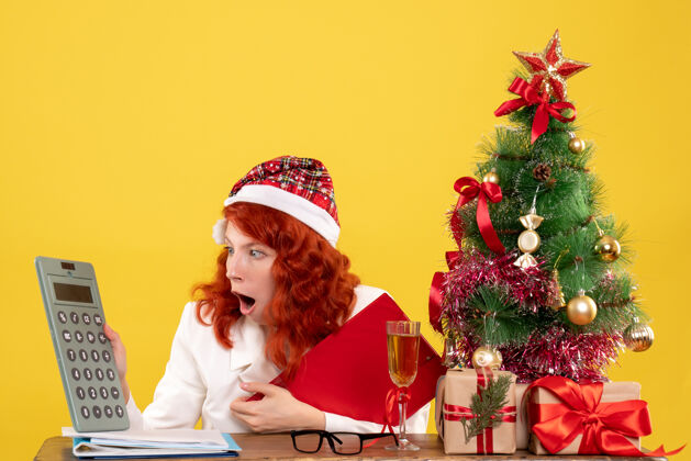 人前视图女医生坐在桌子后面拿着礼物拿着黄色背景上的计算器 还有圣诞树和礼品盒计算器桌子礼物