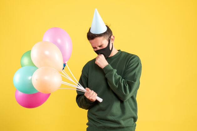乐趣正面图：戴着派对帽 站在黄色地面上的彩色气球的忧心忡忡的年轻人五颜六色生日帽子