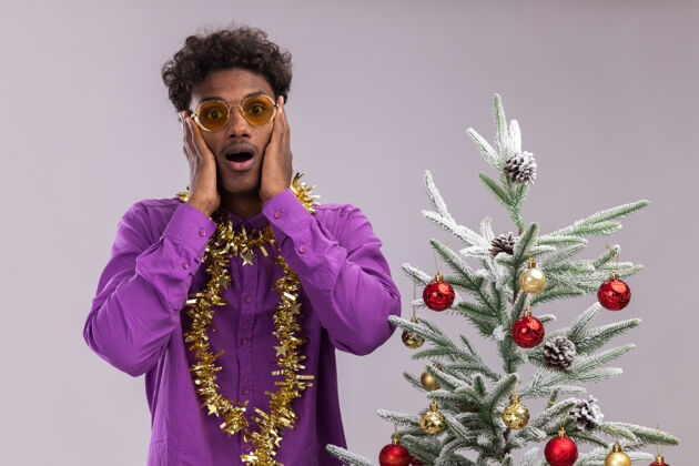 金箔令人印象深刻的年轻黑人男子戴着眼镜 脖子上戴着金属丝花环 站在装饰过的圣诞树旁 手放在脸上 隔离在白色的墙上脖子站着年轻人