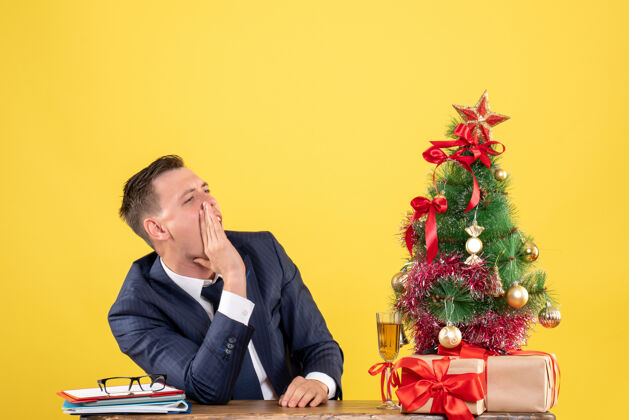 树帅哥打电话给坐在圣诞树旁桌子旁的人的正面图 黄色的礼物电话西装主任