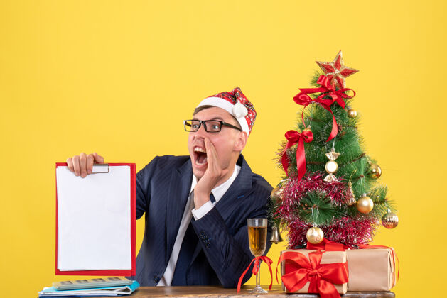男人前视图：手持剪贴板的商人坐在圣诞树旁的桌子旁 黄色的礼物男性办公室桌子