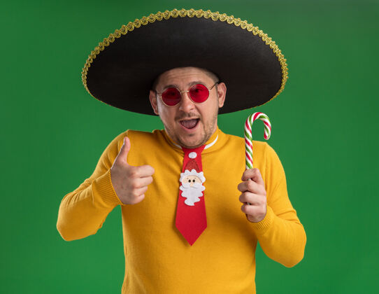 圣诞节穿着黄色高领毛衣 戴着眼镜 戴着滑稽的红色领带 戴着黑色帽子 手里拿着糖果手杖 脸上带着幸福的笑容 竖起大拇指站在绿色的墙上年轻男人糖果