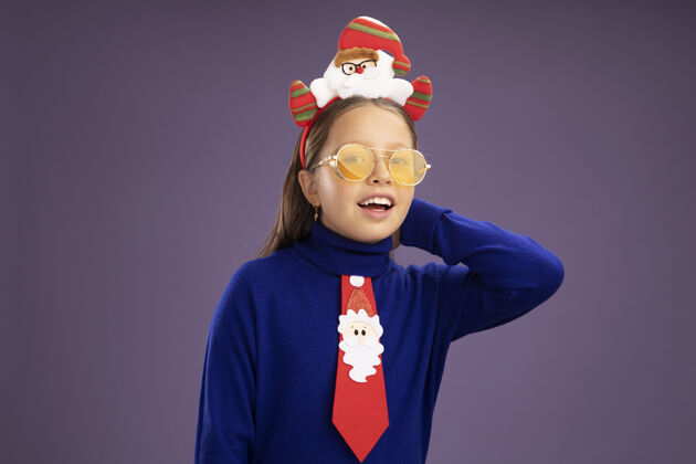 头小女孩穿着蓝色高领毛衣 系着红色领带 头上戴着有趣的圣诞戒指 站在紫色的背景下 看着镜头 脸上洋溢着幸福的笑容搞笑边缘高领毛衣