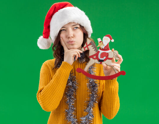 斯拉夫体贴的年轻斯拉夫女孩戴着圣诞帽 脖子上戴着花环 把手指放在下巴上 把圣诞老人抱在摇马装饰上 看着绿色背景上孤立的一面 还有复制空间摇摆圣诞老人下巴