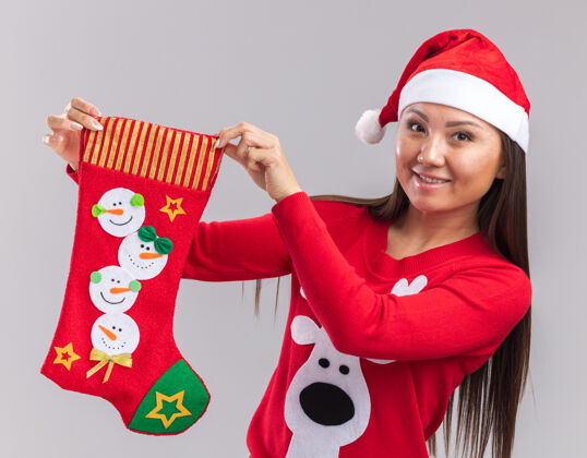 帽子笑容满面的亚洲女孩戴着圣诞帽 穿着毛衣 手里拿着圣诞袜 孤零零地站在白色的墙上圣诞节女人年轻人