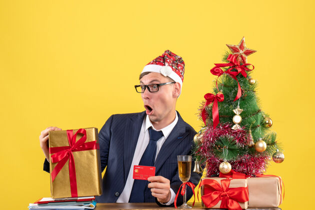 黄色前视图震惊的男子拿着卡片坐在圣诞树附近的桌子上 黄色的礼物职业新郎圣诞节