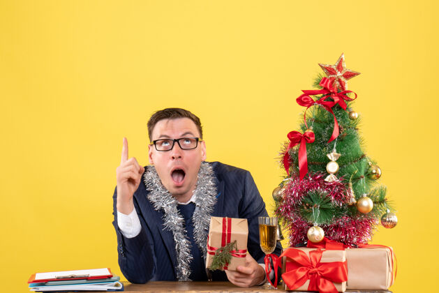 肖像令人惊讶的男子手指指着坐在圣诞树附近的桌子和黄色墙上的礼物前视图坐着人前面