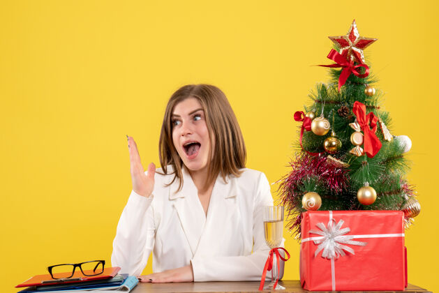 前面正面图：女医生坐在黄色背景的桌子前 放着圣诞树和礼品盒专业人圣诞节