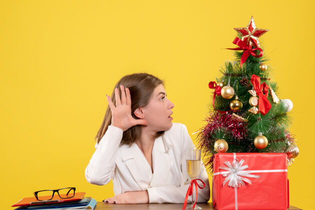 快乐正面图：女医生坐在黄色背景的桌子前 放着圣诞树和礼品盒人圣诞节男人
