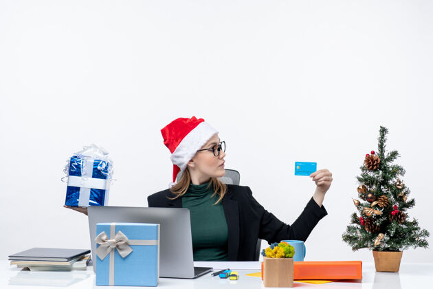 圣诞老人戴着圣诞老人帽戴着眼镜的女商人坐在桌子旁 手里拿着圣诞礼物和银行卡树快乐商务女人