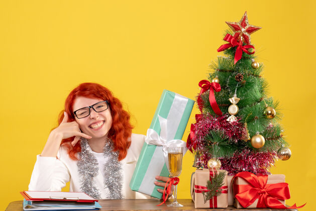 女正面图女医生坐在黄色的桌子上 手里拿着圣诞礼物和圣诞树前面新郎快乐