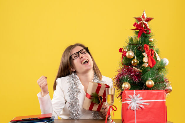 女医生正面图：女医生坐在桌子前 拿着黄色背景的礼物 还有圣诞树和礼品盒健康现在女性