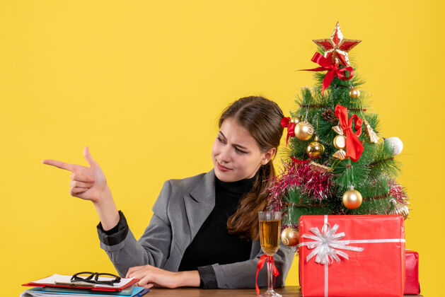 满意正面图满意的女孩坐在桌旁用手指着圣诞树旁的东西和礼物鸡尾酒鸡尾酒女孩礼物