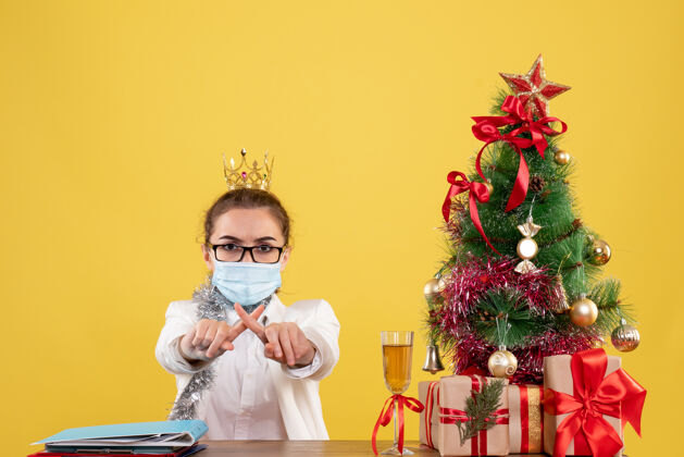 人正面图女医生戴着无菌口罩坐在黄色背景上 带着圣诞树和礼品盒快乐不育脸