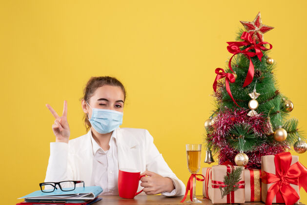 医生正面图女医生戴着防护面具坐在黄色背景上 带着圣诞树和礼品盒情绪背景男性