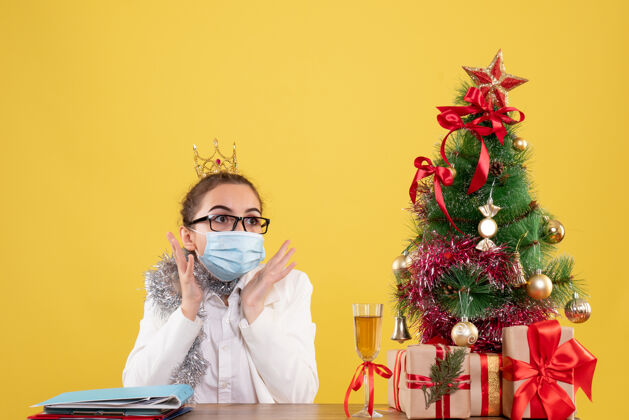 背景正面图女医生戴着黄色背景的无菌口罩坐在圣诞树和礼盒里前面漂亮脸