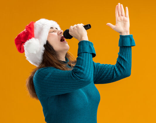 圣诞老人戴着圣诞帽的成年白种女人拿着麦克风 假装在橙色背景上用复制空间孤立地唱歌快乐假装帽子