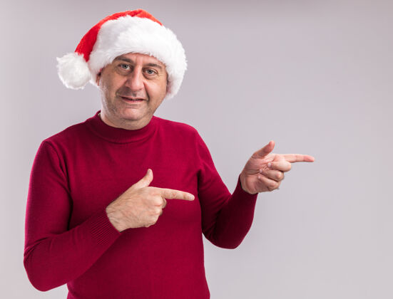 圣诞老人戴着圣诞老人帽的中年男子面带微笑地看着摄像机 站在白色背景上 用手指着旁边的五线谱年龄站着相机