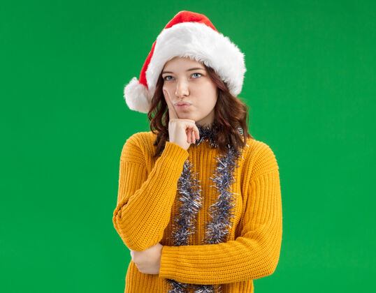 看体贴的年轻斯拉夫女孩戴着圣诞帽 脖子上戴着花环 把手指放在脸上 看着绿色背景上孤立的一面 留着复制空间圣诞节年背景