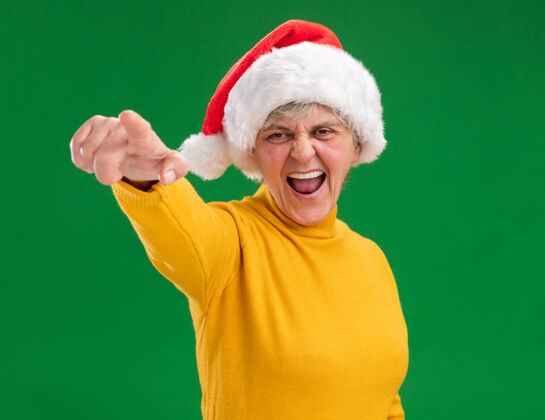 看戴着圣诞帽的恼怒的老妇人看着并指着隔离在紫色背景上的相机 还有复印空间年相机背景