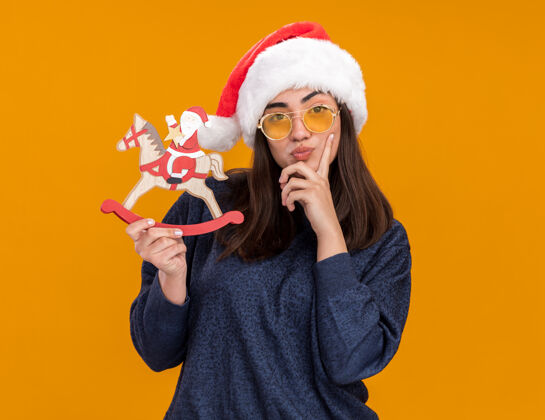 快乐体贴的年轻白种女孩戴着太阳眼镜 戴着圣诞老人帽 手指放在下巴上 把圣诞老人抱在摇马上 橙色背景上有复制空间年放手指
