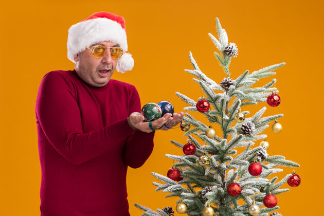 年龄中年男子戴着圣诞老人帽 穿着深红色高领毛衣 戴着黄色眼镜 手里拿着圣诞球 站在橙色背景下的圣诞树旁 看上去很惊讶背景帽子圣诞快乐