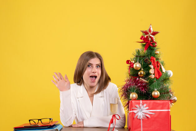 女性前视图女医生坐在桌子前 带着圣诞礼物和黄色背景的树医生人快乐