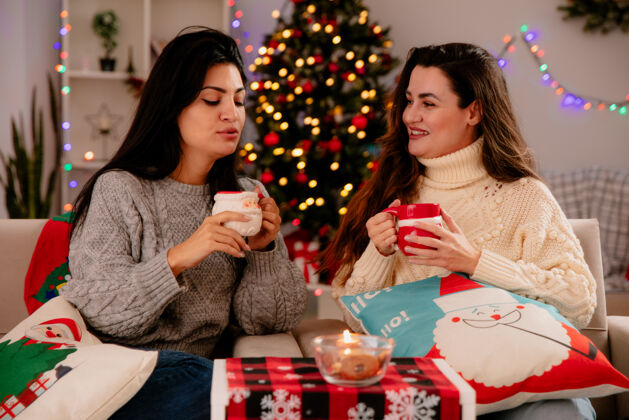 杯子微笑的年轻女孩拿着杯子 看着她高兴的朋友拿着杯子坐在扶手椅上闭着眼睛 在家里享受圣诞节的时光圣诞节抱着微笑