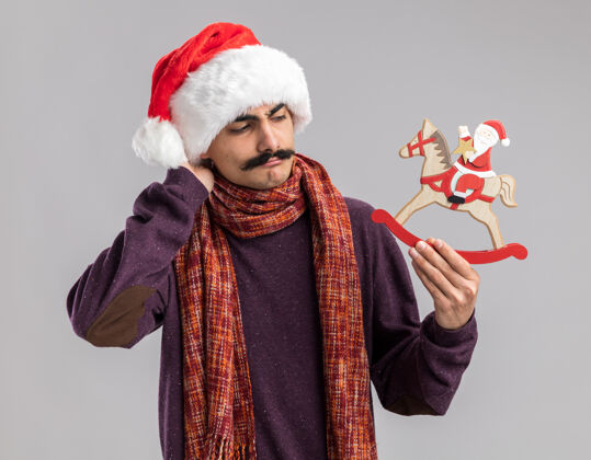 年轻小胡子男人戴着圣诞老人帽 脖子上围着暖和的围巾 手里拿着圣诞玩具 站在白色的背景下困惑而焦急地看着它站着围着圣诞快乐