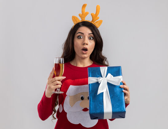 圣诞快乐印象深刻的年轻漂亮女孩穿着驯鹿鹿角头带和圣诞老人毛衣拿着香槟和礼品包看新年年轻礼物