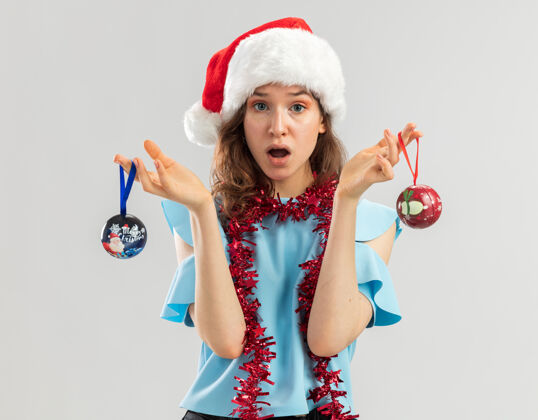 圣诞快乐身穿蓝色上衣 戴着圣诞帽 脖子上戴着金属丝的年轻女子拿着圣诞球 看上去既困惑又惊讶周围看女人
