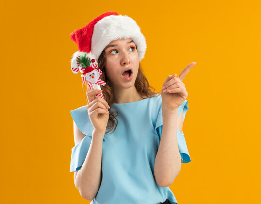 女人身穿蓝色上衣 戴圣诞帽的年轻女子手持圣诞糖果手杖 惊讶地用食指指着什么东西东西指着手指