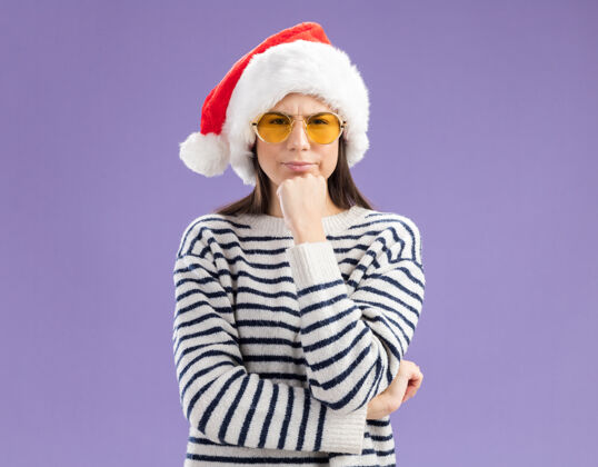 快乐戴着太阳眼镜 戴着圣诞帽的困惑的白人女孩托着下巴 神色很好年轻下巴圣诞