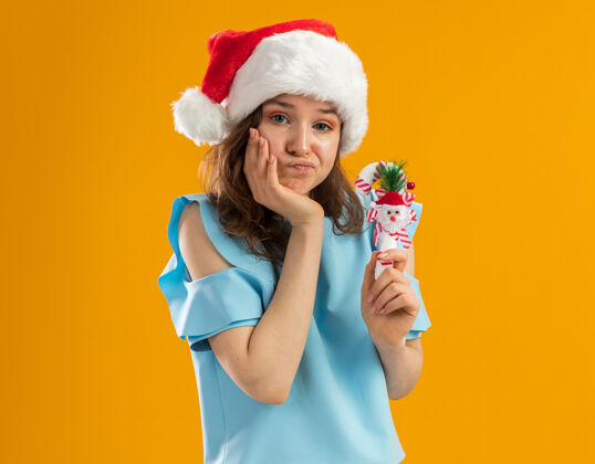 蓝色身穿蓝色上衣 戴圣诞帽 手持圣诞糖果手杖的年轻女子 手放在下巴上 看上去既困惑又不高兴年年轻不高兴