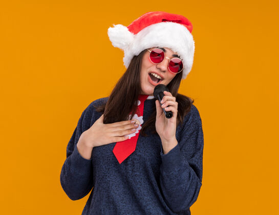 帽子戴着太阳眼镜 戴着圣诞帽 系着圣诞领带的年轻白人女孩高兴地把手放在胸前 拿着麦克风假装唱歌年胸部复制