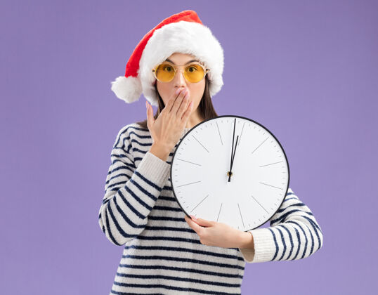 时钟一个戴着太阳眼镜 戴着圣诞帽的年轻白人女孩惊讶地把手放在嘴上 手里拿着钟空间女孩紫色