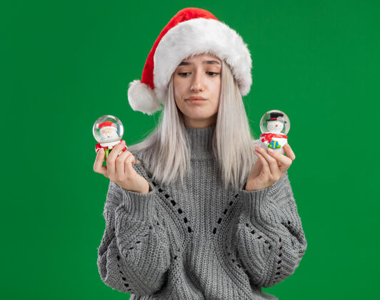 圣诞快乐年轻的金发女郎穿着冬衣 戴着圣诞帽 手里拿着圣诞玩具雪球 困惑地看着相机 站在绿色的背景下做着选择站着帽子毛衣